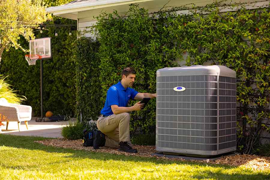 A technician in a blue shirt crouches beside an outdoor air conditioning unit, using tools. Lush greenery and a basketball hoop are in the background.