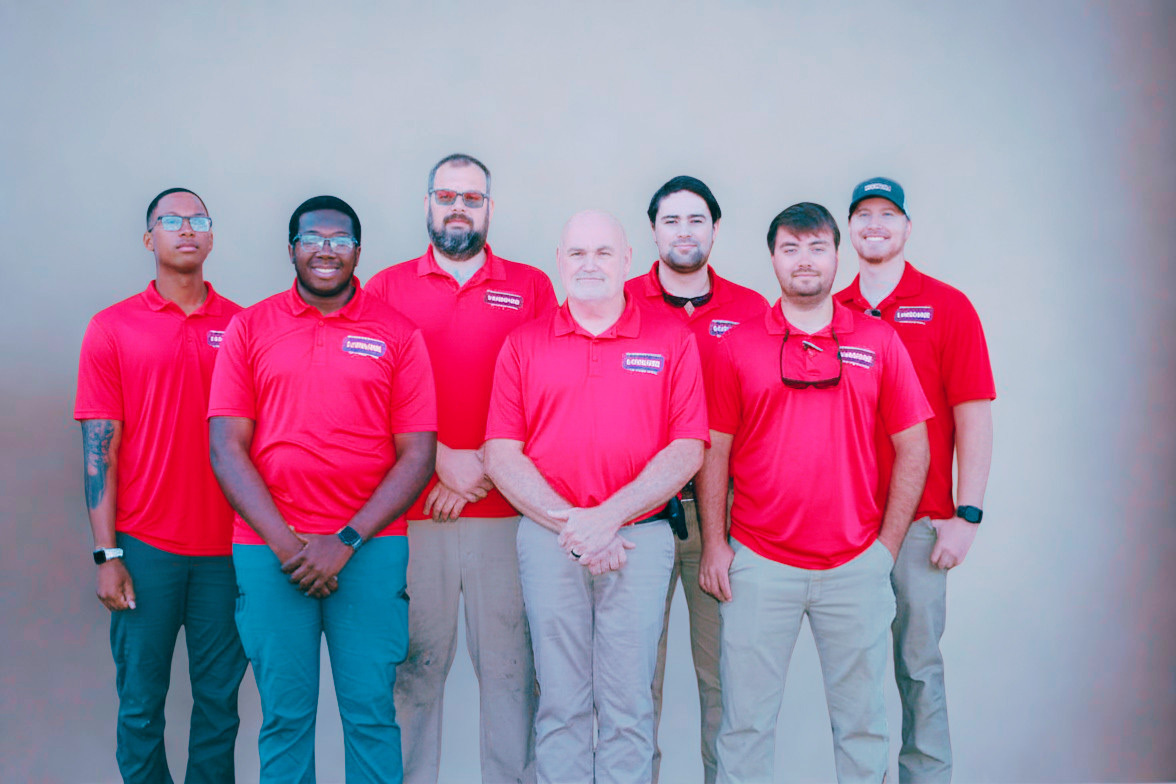 A group of seven men wearing red polo shirts standing in front of a neutral background.
