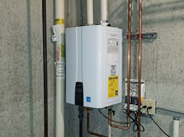 A white tankless water heater mounted on a concrete wall, surrounded by copper pipes and an electrical conduit. The setting is a dimly lit utility room.