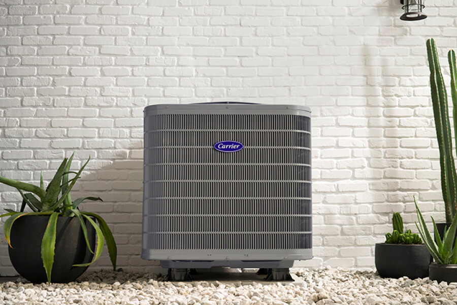 An outdoor air conditioner unit with a "Carrier" logo, set against a white brick wall. Surrounded by green plants in black pots on white gravel.