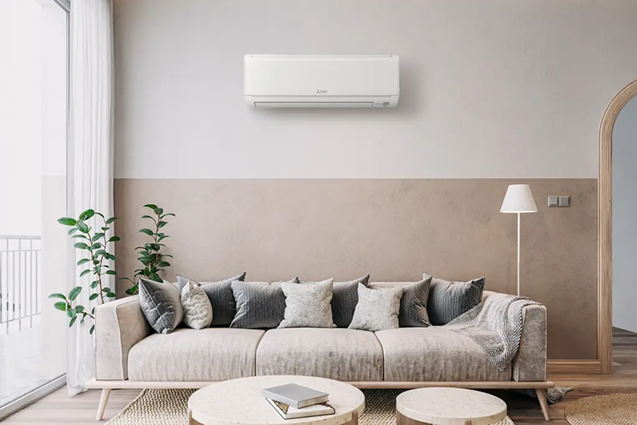 A modern living room with a beige and cream color scheme, featuring a light-colored sofa adorned with decorative pillows, a floor lamp, and indoor plants. An air conditioning unit is mounted on the wall above the sofa.